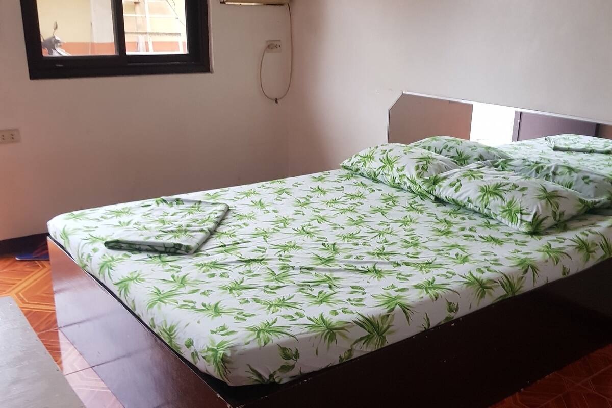 Un lit simple orné d’un motif de feuilles vertes, une petite table et une fenêtre donnant sur l’extérieur.