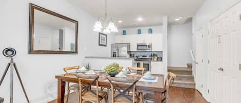 Un comedor con una mesa y sillas de madera, una araña de techo y una cocina al fondo.