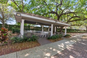 A covered outdoor seating area with tables and chairs, surrounded by trees and a brick pathway. - Your VIP Home near Disney! (Osceola County)