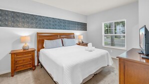 A bedroom with a bed, two bedside tables with lamps, a television, and a window with blinds. - Your VIP Home near Disney! (Osceola County)