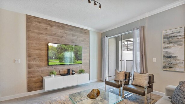 A modern living room with a flat-screen TV mounted on a wooden wall, a glass coffee table, a sofa, and a large window with curtains. - Outstanding 5BDR Disney Vacation (Kissimmee)