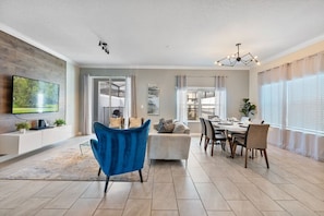 A modern living room with a flat-screen TV, a blue armchair, a dining area with a table and chairs, and a kitchen visible in the background. - Outstanding 5BDR Disney Vacation (Kissimmee)