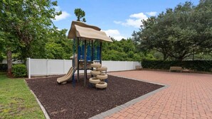 Un parque infantil con tobogán y estructura para escalar, rodeado por un sendero de ladrillo y follaje.