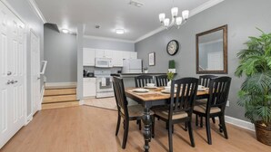 Un comedor con una mesa y sillas de madera, una cocina al fondo, un reloj y una planta en maceta.