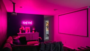 Una habitación con una pared oscura, un letrero de neón que dice "Woof Woof", un proyector, un armario blanco y una pantalla grande.