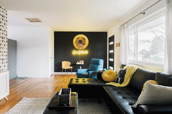 Uma sala de estar moderna com um sofá de couro preto, uma poltrona azul e um banquinho preto. Um letreiro de neon com a imagem de um cachorro e o texto “Life is a Collection” é visível na parede.
