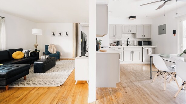 Una sala de estar moderna con un sofá negro, una mesa de centro blanca y un reposapiés negro. Hay un televisor sobre un mueble bajo, una lámpara de pie y una pequeña mesita de noche con un jarrón. La cocina tiene armarios blancos, un microondas y un comedor con una mesa y sillas blancas.