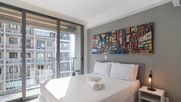 A modern hotel room with a large bed, a nightstand with a lamp, and a colorful abstract painting on the wall. - Studio On Imarés Moema (São Paulo)