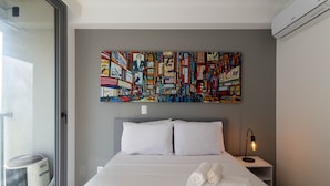 A hotel room with a large bed, white linens, a headboard, a nightstand with a lamp, and a colorful abstract painting on the wall. - Studio On Imarés Moema (São Paulo)