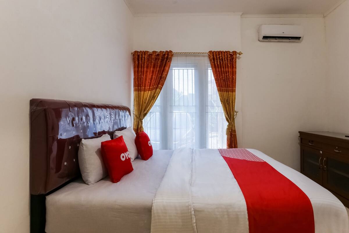 A neatly made bed with a white and red bedspread, two red pillows, and a brown headboard. There is a window with curtains, an air conditioning unit on the wall, and a wooden cabinet to the right.
