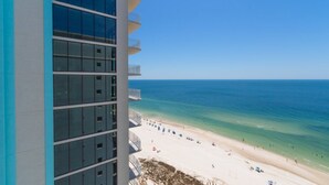 Um hotel alto com vista para a praia, com varandas e guarda-corpos, e uma praia arenosa com guarda-sóis.