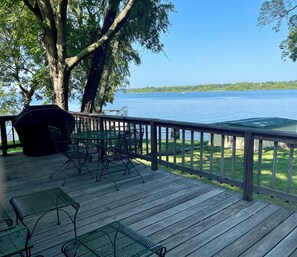 Eine Holzterrasse mit Metallstühlen und einem Grill, mit Blick auf einen stillen See und eine gräserbewachsene Uferlinie.