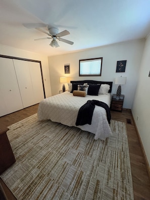 Une chambre à coucher comprenant un lit, un ventilateur de plafond, une table de chevet avec une lampe, une fenêtre avec des stores et une porte.