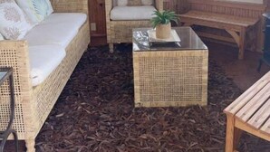 Een houten chalet met een rieten bank, een stoel en een kleine tafel met een plant. Er zijn ramen met uitzicht op een meer en boten.