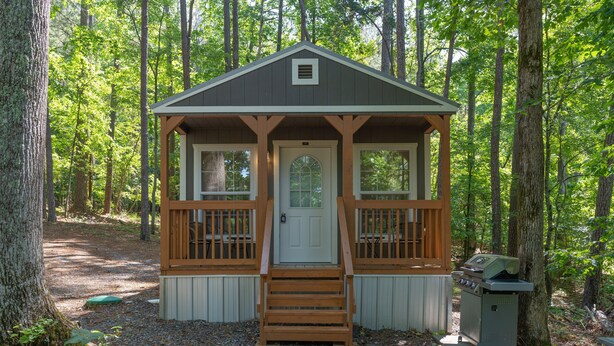 A small wooden cabin with a front door, a porch with railings, and a grill outside.
