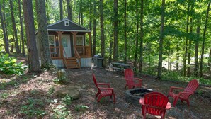 A cabin with a deck, surrounded by trees, and a fire pit with chairs.