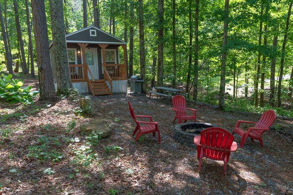 A cabin with a deck, surrounded by trees, and a fire pit with chairs.
