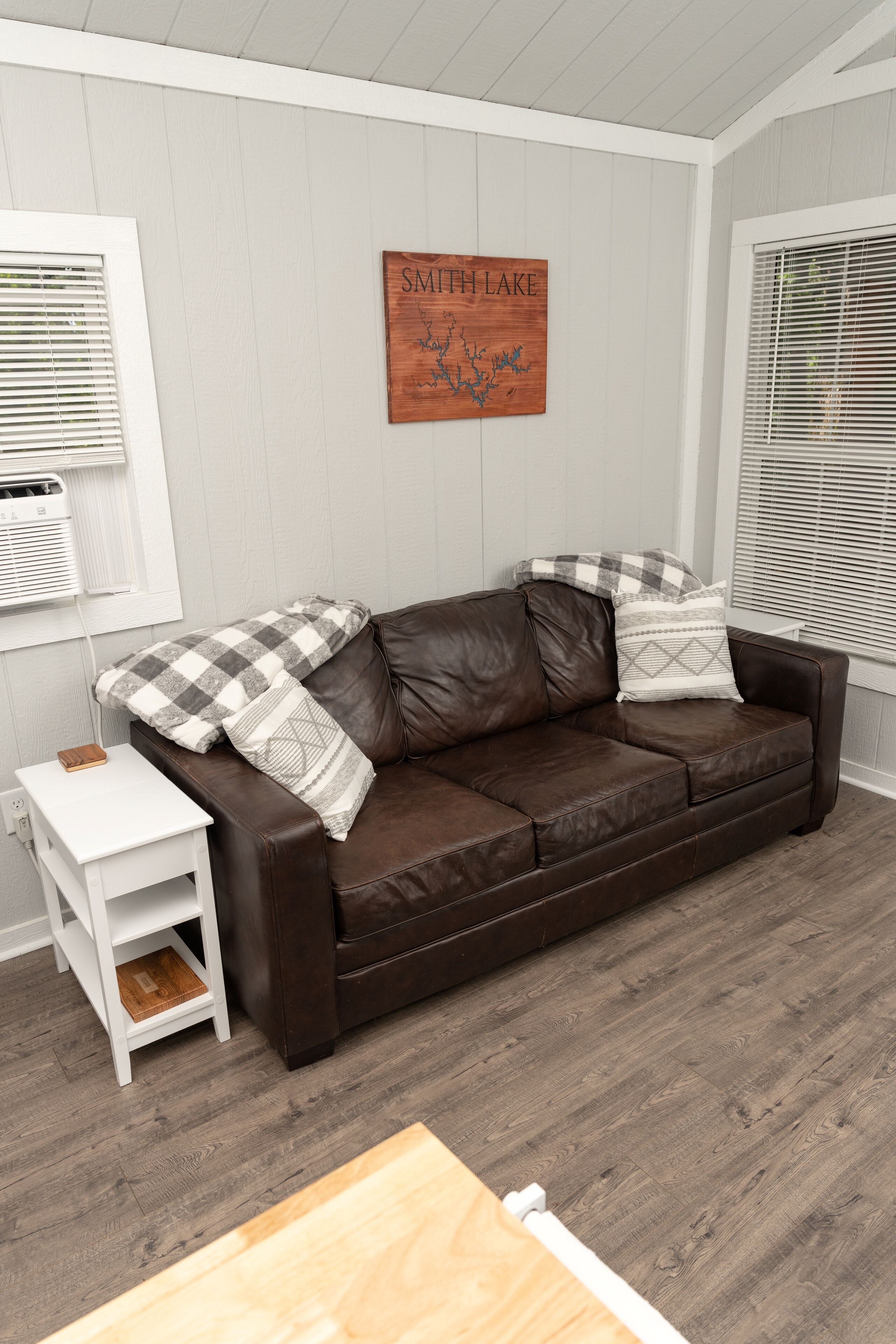 A brown leather sofa with checkered and patterned pillows, a small white side table, and a wooden sign on the wall.