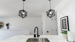 Une cuisine moderne dotée d’un plan de travail blanc, d’un évier en acier inoxydable, d’un robinet noir et de deux suspensions géométriques noires.