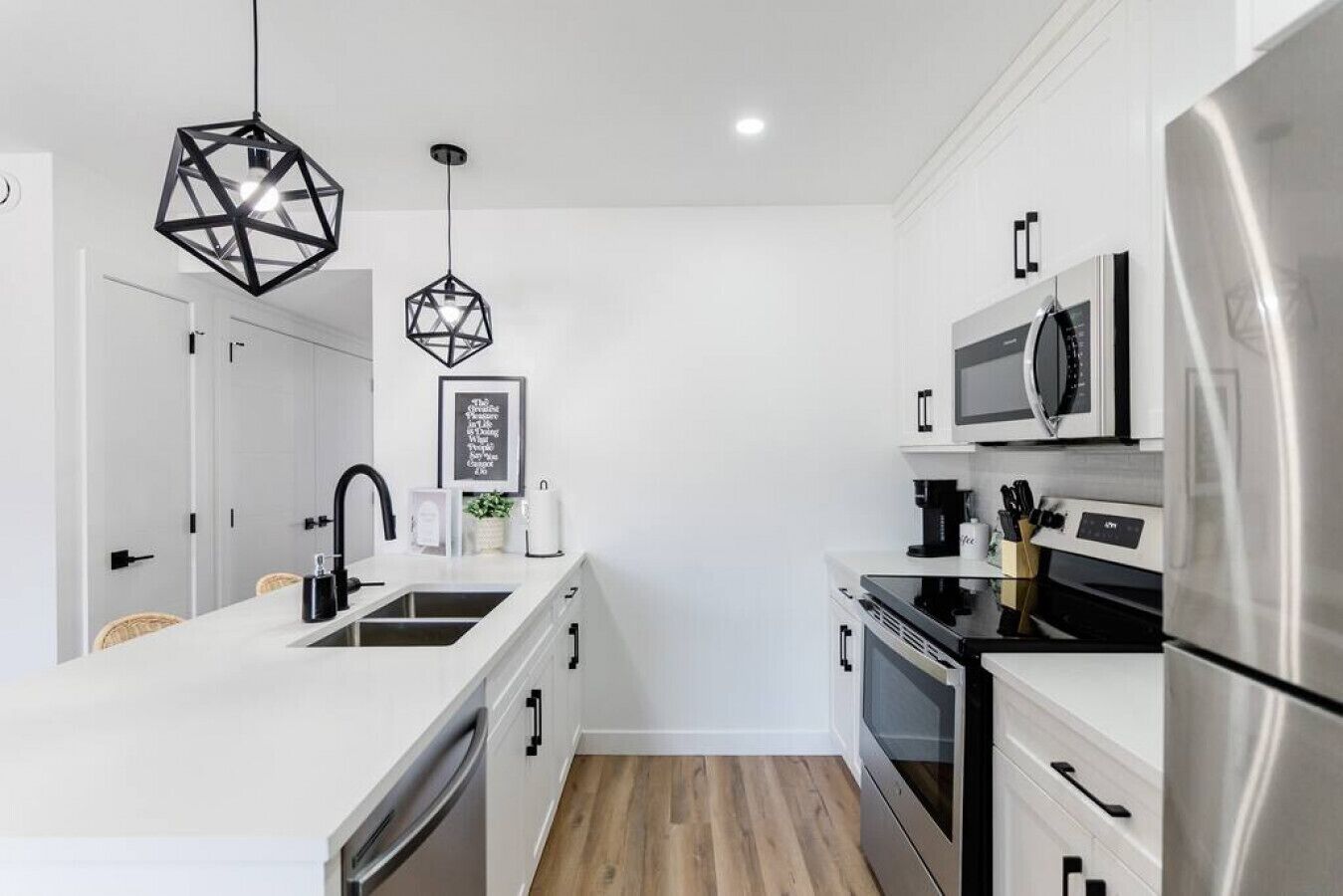 A modern kitchen with white cabinetry, stainless steel appliances, a central island with a sink, and geometric pendant lighting.