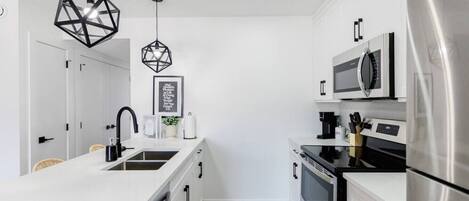 A modern kitchen with white cabinetry, stainless steel appliances, a central island with a sink, and geometric pendant lighting.