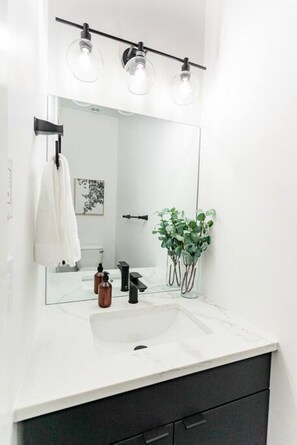 Une salle de bain moderne avec un lavabo blanc, des meubles sombres et un grand miroir agrémenté de trois luminaires.