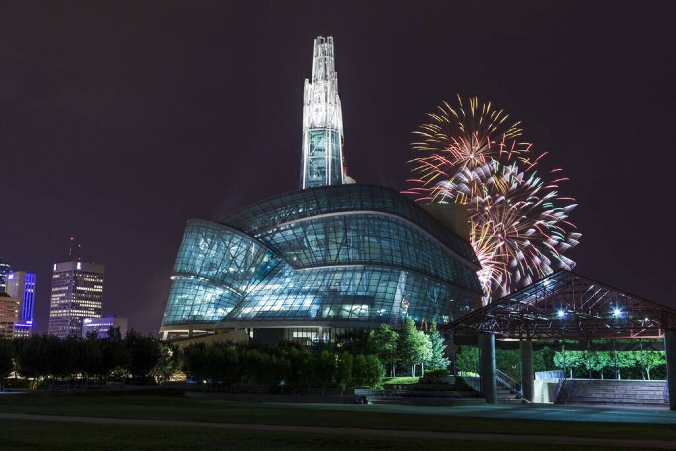 A modern glass building with a unique curved design and a tall spire, surrounded by a park area and fireworks display.