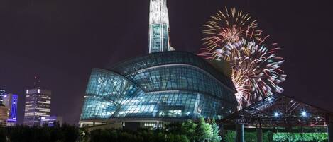 A modern glass building with a unique curved design and a tall spire, surrounded by a park area and fireworks display.