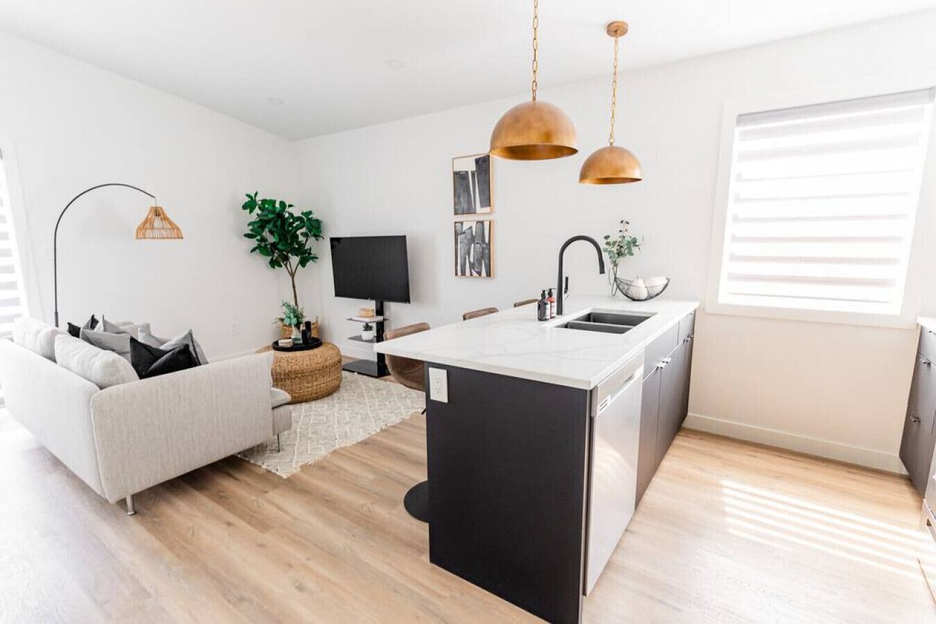 A modern kitchen with a central island, black cabinets, and a white countertop. There is a dining area with a white sofa and a small side table. A large window provides natural light, and a plant adds greenery to the space.
