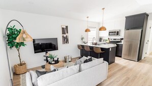 A modern living room with a sofa, a coffee table, and a TV. There is a kitchen area with a fridge, microwave, and bar stools. - Riverview Health | Trendy Area | Free Parking (Winnipeg)