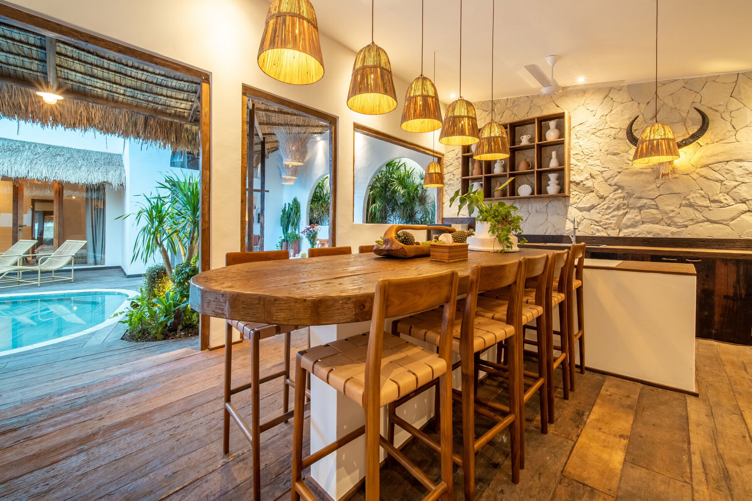 Une table à manger en bois avec des chaises, un coin cuisine avec un mur en pierre et des luminaires suspendus, et une piscine visible à travers une porte vitrée.