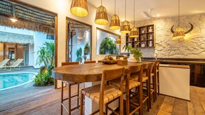Une table à manger en bois avec des chaises, un coin cuisine avec un mur en pierre et des luminaires suspendus, et une piscine visible à travers une porte vitrée.