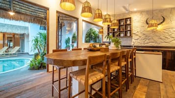 Une table à manger en bois avec des chaises, un coin cuisine avec un mur en pierre et des luminaires suspendus, et une piscine visible à travers une porte vitrée.