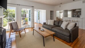 Un salon moderne avec un canapé, une table basse et un fauteuil. Il y a une grande fenêtre avec des rideaux, une vue sur l’extérieur et un coin cuisine avec des étagères.