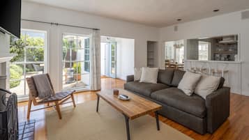 A modern living room with a sofa, coffee table, and a chair. There is a large window with curtains, a view of the outdoors, and a kitchen area with shelves.
