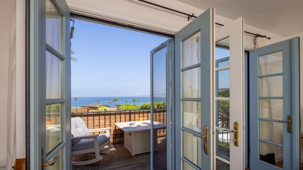 Une porte ouverte donnant sur un balcon avec vue sur l’océan, un plancher en bois et un fauteuil à bascule blanc.