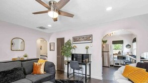 A modern living room with a grey sofa, a black rug, and a ceiling fan. There is a dining area with a black table and chairs, and a kitchen visible in the background. - Shocker Shack - 2K - Near WSU, Park - Fenced Yard (Wichita)