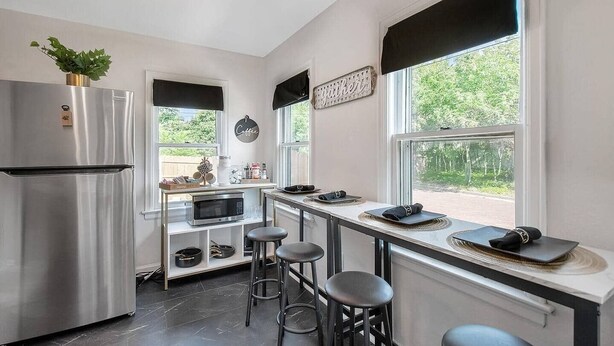 A modern kitchen with a stainless steel refrigerator, a microwave, and a coffee station. There are three stools and a window with a view of greenery. - Shocker Shack - 2K - Near WSU, Park - Fenced Yard (Wichita)