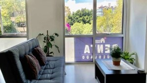 A modern living room with a dark sofa, a small side table with a plant, and large windows offering a view of greenery and buildings.