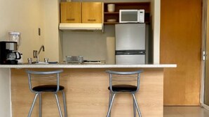 A compact kitchen with a white countertop, two bar stools, a refrigerator, and a microwave.