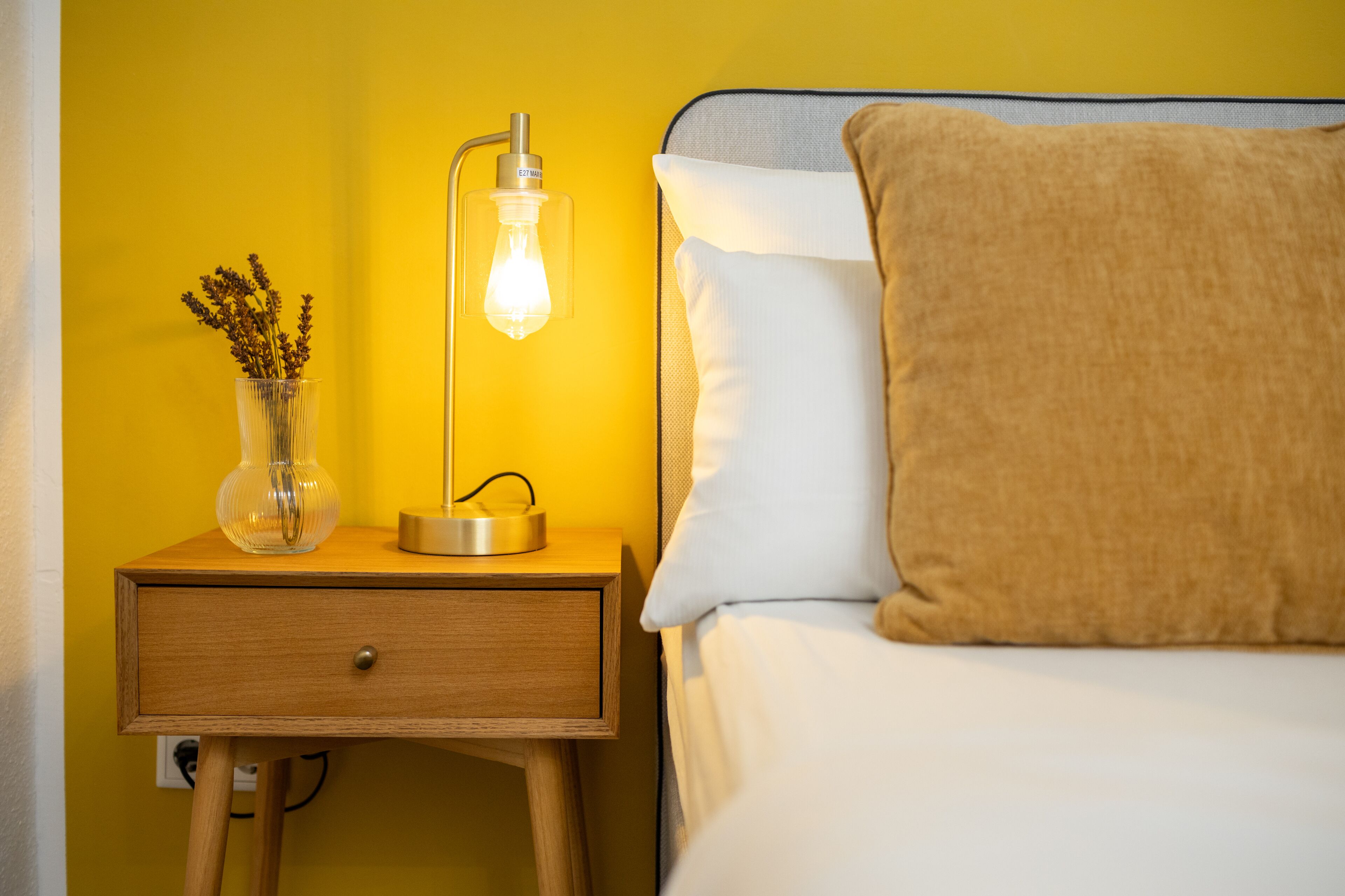 A bed with a white pillow and a brown throw, a bedside table with a lamp, and a vase with dried flowers.