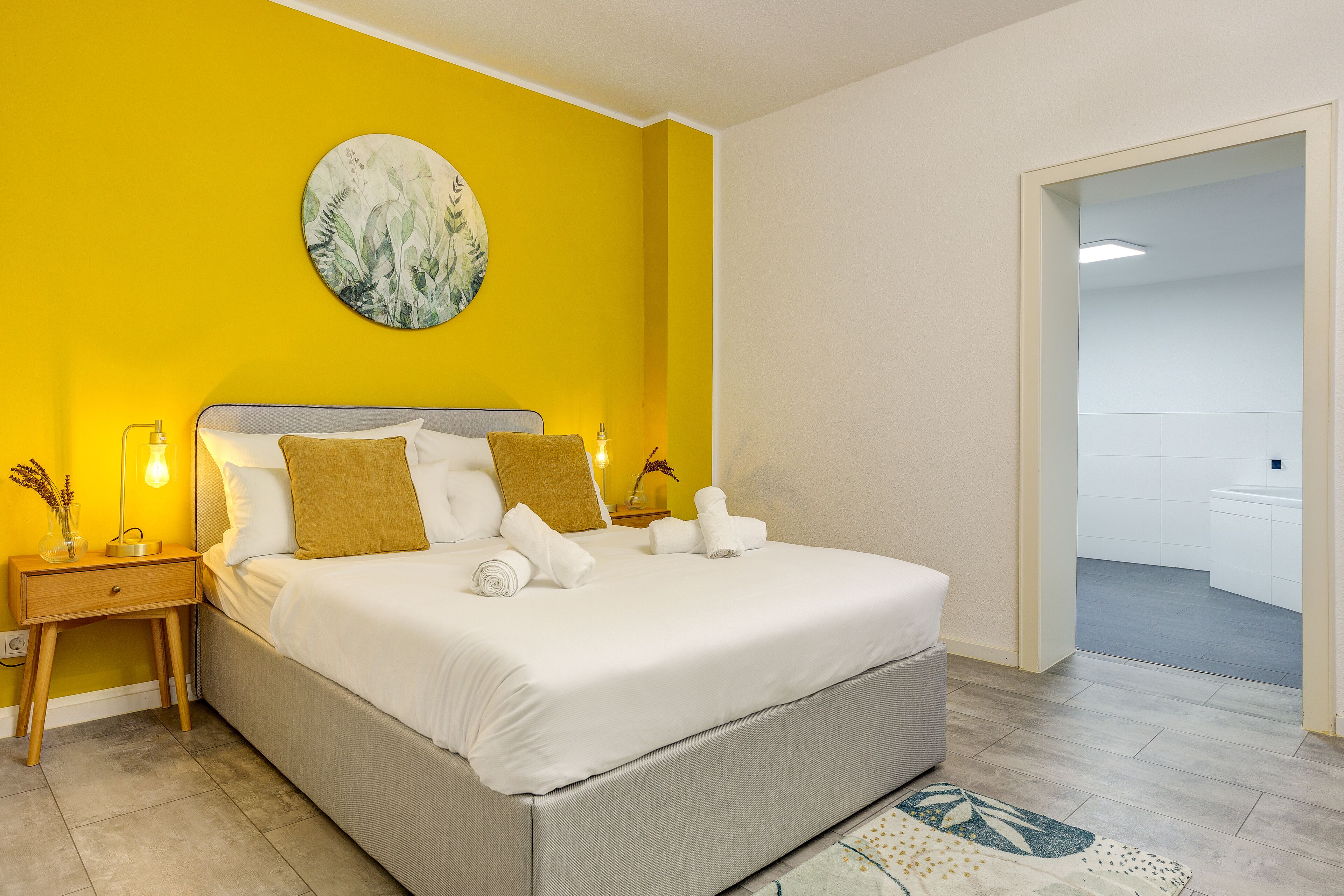 Camera da letto con un letto, un comodino, un'opera d'arte a parete e un bagno visibile attraverso una porta aperta.