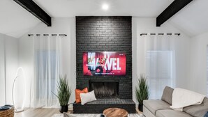 A modern living room with a black brick fireplace, a flat-screen TV mounted above it, a beige sofa, and a coffee table. - Ultimate Comfort & Modern Elegance in our Renovated Pool House with Hot Tub (Carrollton)