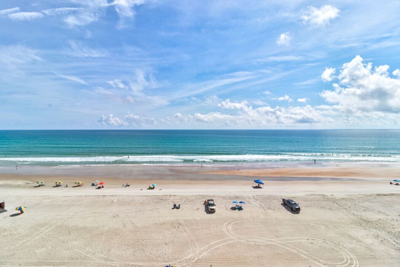 Une plage avec un ciel dégagé, une mer calme, et quelques parasols et véhicules sur le sable.