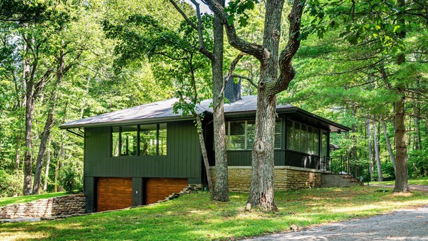 A modern house with a stone foundation, large windows, and a garage, surrounded by a wooded area.