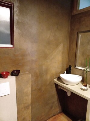 Une salle de bain avec un lavabo blanc, deux bols rouges et un bol brun foncé posés sur le comptoir, un miroir au-dessus du lavabo et une fenêtre donnant sur le ciel.