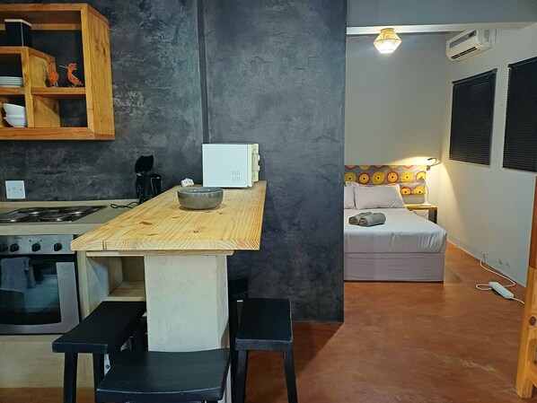 Une cuisine compacte avec un plan de travail en bois, un micro-ondes blanc et une cuisinière. On y trouve un petit coin repas avec deux chaises noires et un bol posé sur le plan de travail. La pièce est équipée d’un lit avec une tête de lit colorée et d’une table de chevet avec une lampe.