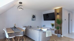 Ein modernes Wohnzimmer mit einer Couch, einem Esstisch und Stühlen. An der Wand ist ein Fernseher angebracht und es steht ein Topfpflanze dort.