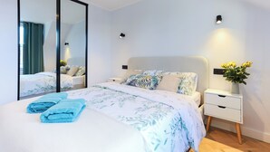Ein modernes Schlafzimmer mit einem großen Bett, einem Nachttisch mit Blumenvase und einem Spiegel, der den Raum reflektiert.