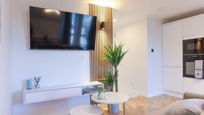 Ein modernes Wohnzimmer mit einem an der Wand befestigten Flachbildfernseher, einem weißen Konsolentisch mit Dekoration und einem runden Couchtisch sowie einem Blumentopf.